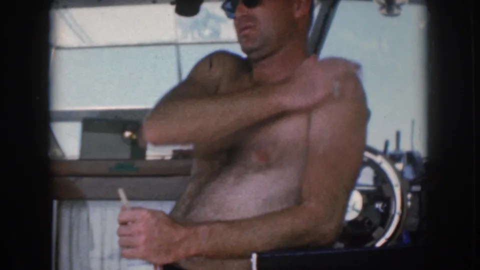 1961 Michigan boating footage frame with a shirtless man in sunglasses adjusting his shoulder near the helm, cigarette still visible.