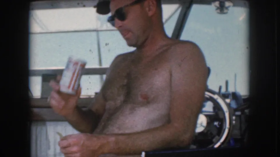 1961 Michigan home-movie frame showing the man at the boat helm holding a beverage can in one hand and a cigarette in the other.