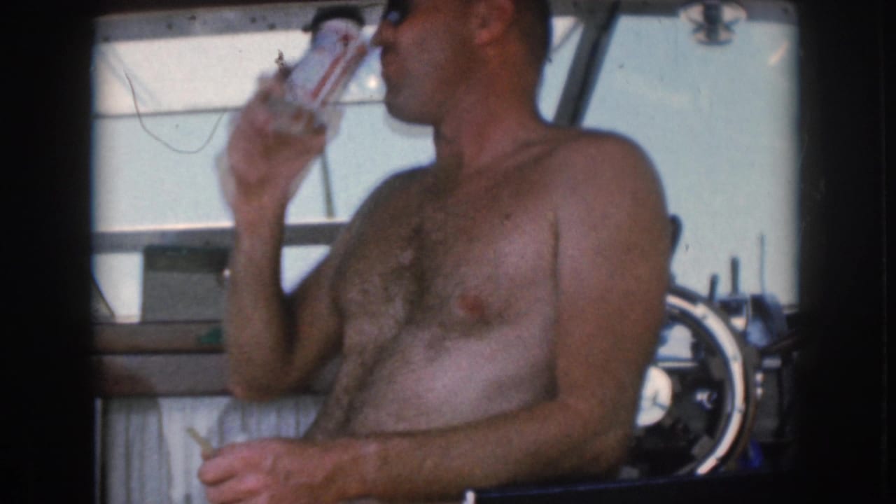 Shirtless man seated on a boat drinks from a can beside the helm.