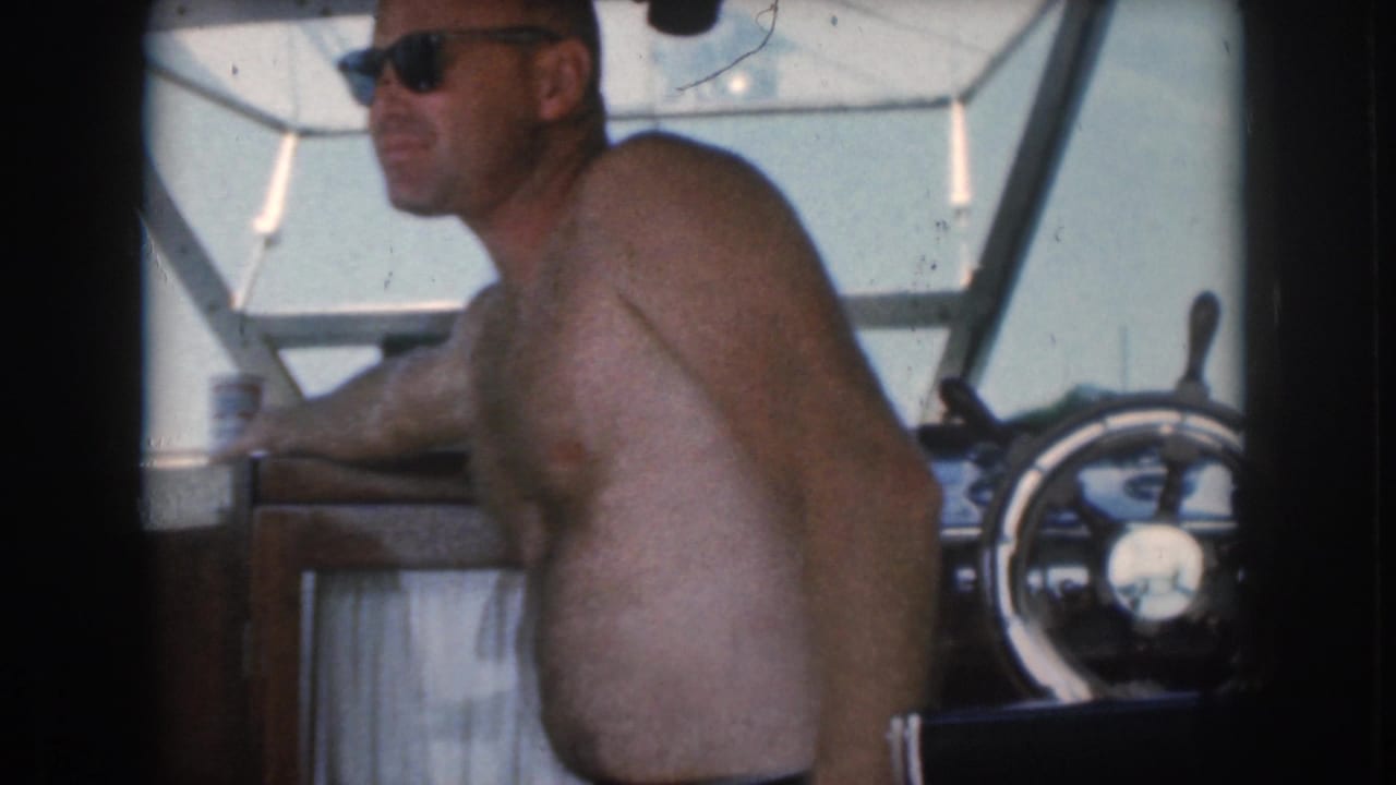Shirtless man in sunglasses sits at the boat helm with a can nearby.