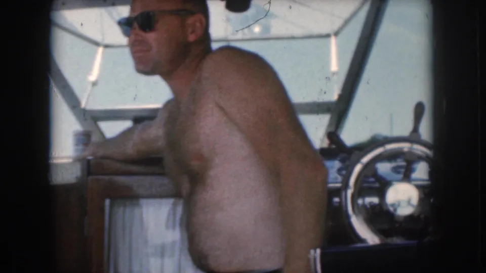 A shirtless man wearing sunglasses relaxes at the helm of a motorboat in the 1961 Grand Rapids, Michigan home-movie scene.