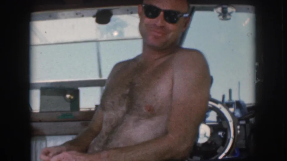 The shirtless man looks toward the camera from his seat inside the boat, with the helm and windshield visible behind him.