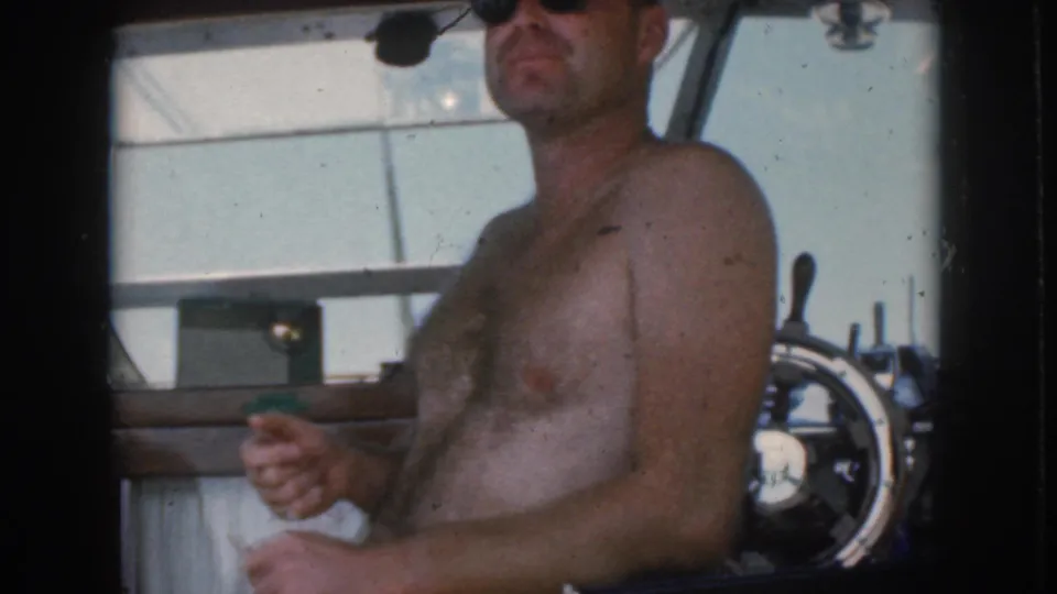 The man remains seated near the motorboat helm, his face partly cropped as he holds one hand up in the 8mm frame.