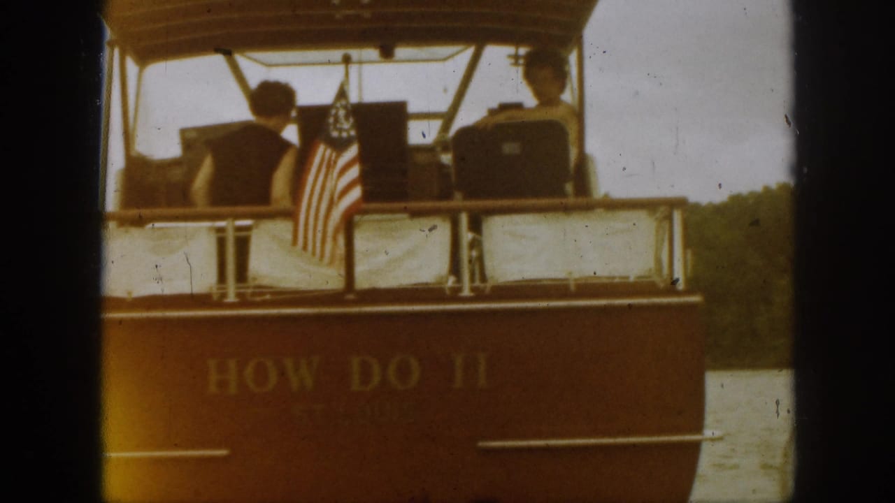 In metadata-supported 1961 Michigan footage, a covered motorboat named How Do II is seen from the stern with two passengers and an American flag.