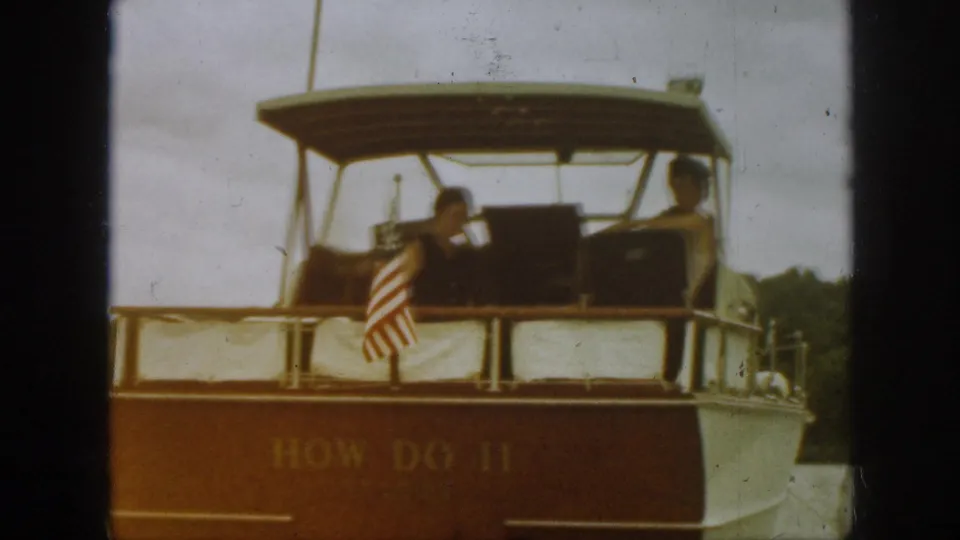 The boat How Do II appears from behind on a Michigan lake, with two seated passengers, an American flag, stern railings, and wooded shoreline.