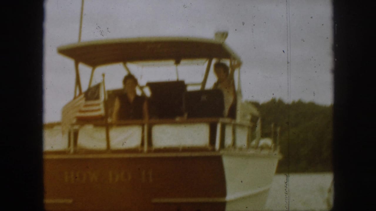1961 Michigan home-movie frame showing a white and brown motorboat on a lake, with two adults visible beneath a canvas canopy.