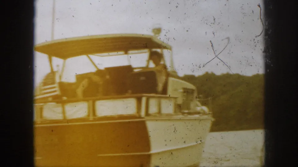 1961 Michigan boating footage frame with a classic motorboat cruising near a wooded shoreline, one adult visible by the canopy.