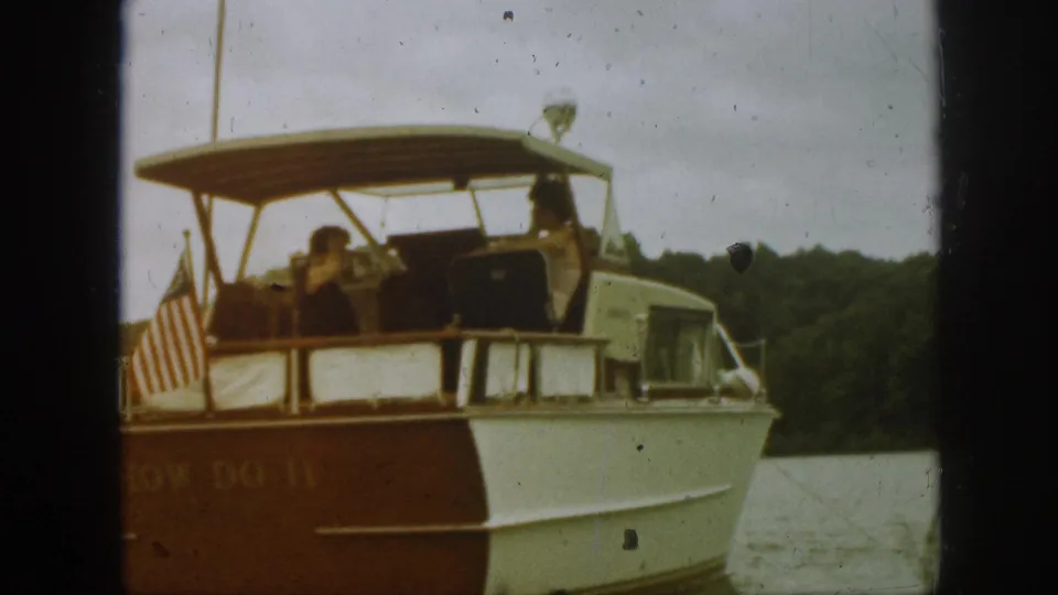 1961 Michigan home-movie frame showing a motorboat with an American flag at the stern and adults aboard on a lake.
