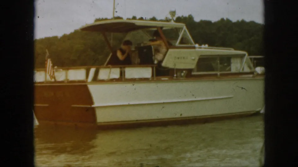 1961 Michigan frame showing the side of a white and brown motorboat on a lake, with adults under the canopy and wooded shore behind.