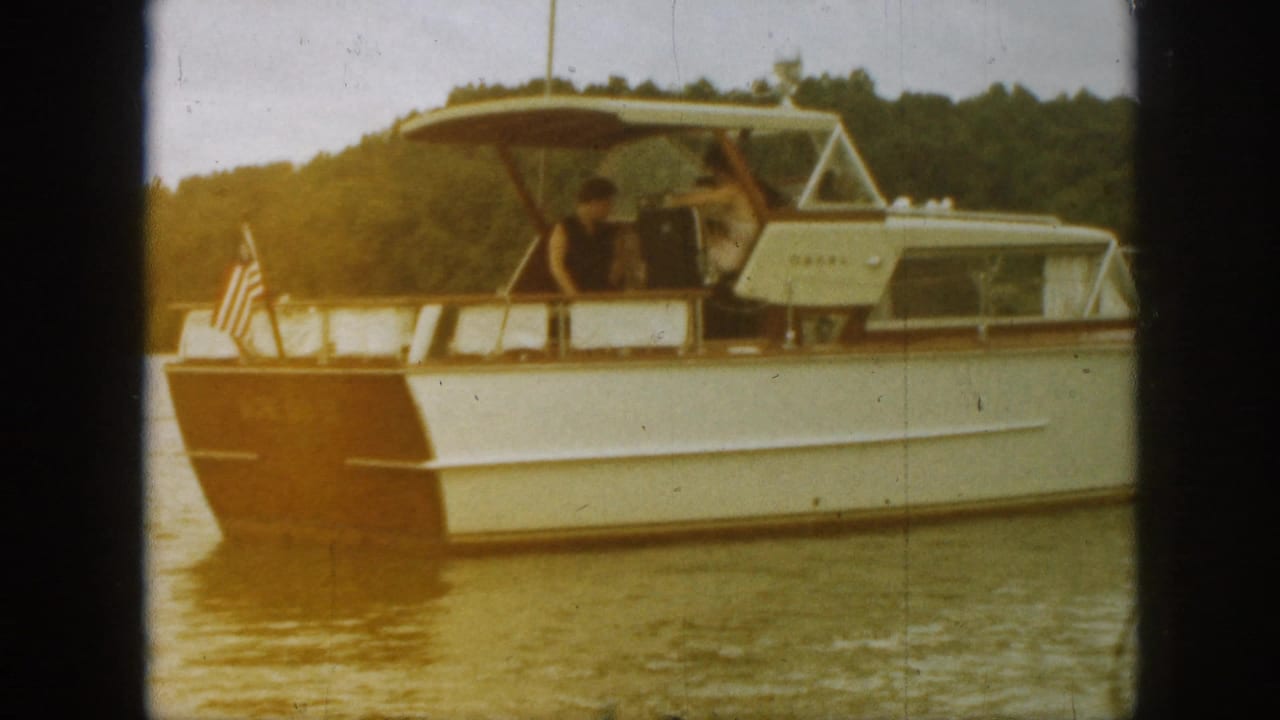 1961 Michigan home-movie frame showing a white and brown cabin cruiser on lake water with people on deck and trees behind it.