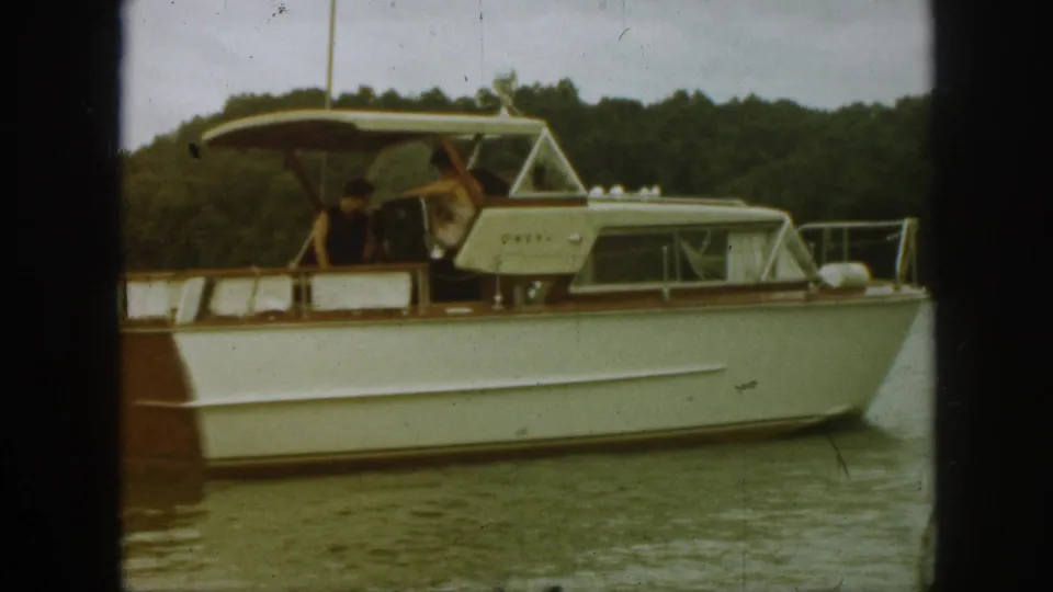 1961 Michigan 8mm frame of a cabin cruiser on calm water, with two visible people near the canopy and wooded shoreline beyond.