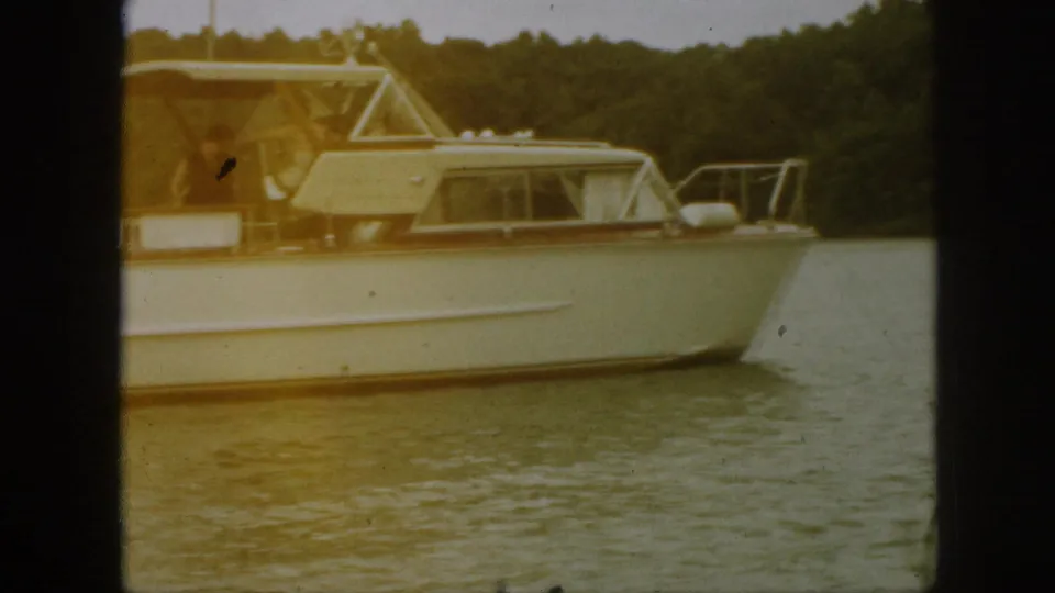 1961 Michigan 8mm frame of a white and brown cabin cruiser on the lake, with a person near the canopy and trees in the background.