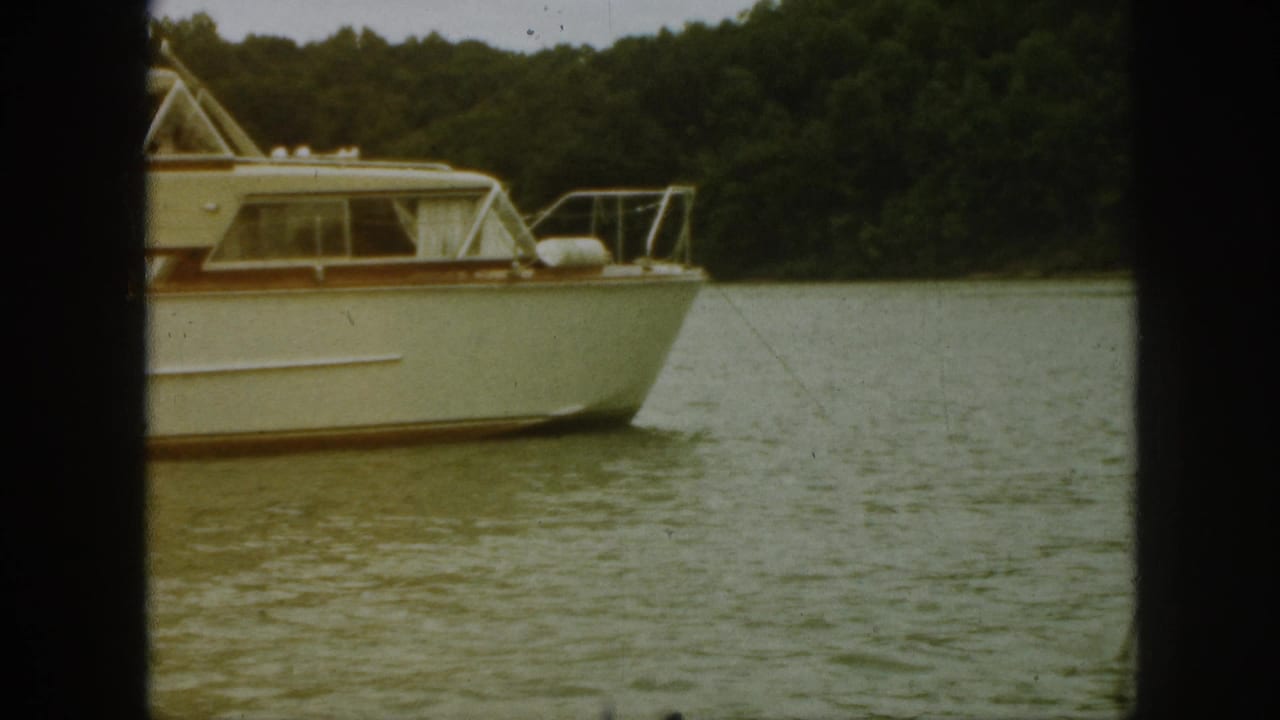 1961 Michigan home-movie frame showing a white cabin cruiser passing on a wooded river.