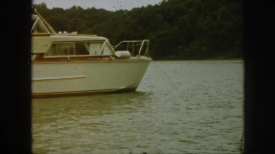 1961 Michigan home-movie frame showing a white cabin cruiser passing on a wooded river.