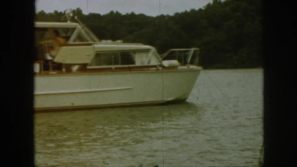 1961 Michigan frame of a white motorboat with wood trim cruising past trees, with a person visible near the helm.