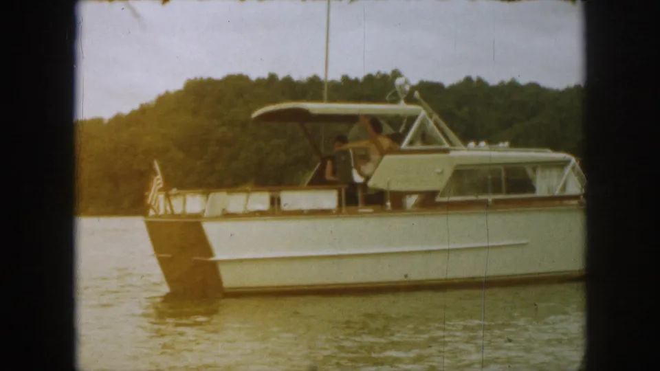1961 Michigan home-movie frame showing the stern of a white cabin cruiser with a small flag against wooded shoreline.