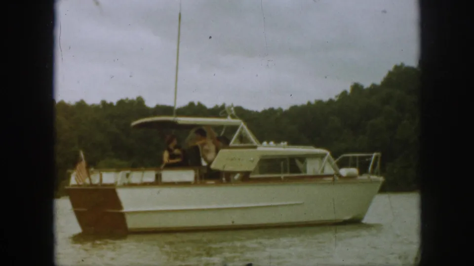 Broadside 1961 Michigan home-movie view of the white motorboat with brown stern, canopy, stern flag, and people aboard.