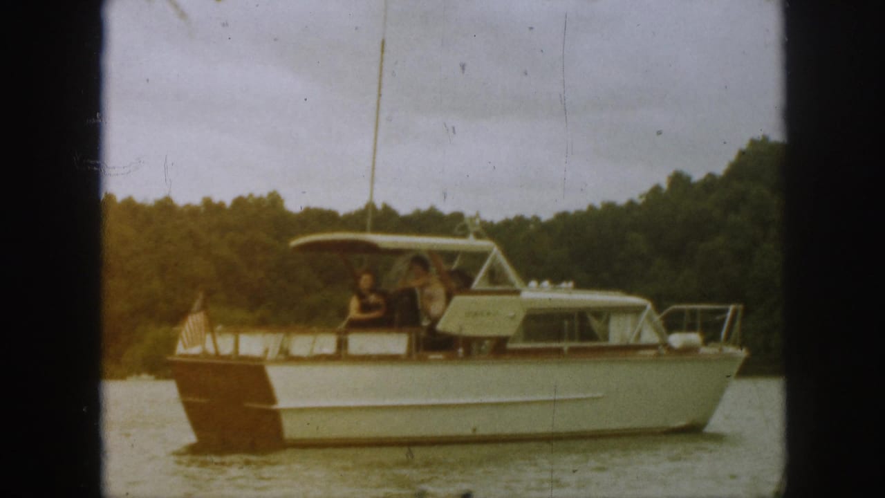 1961 Michigan home-movie frame of a cabin-style motorboat crossing calm lake water with passengers visible under the rear canopy.