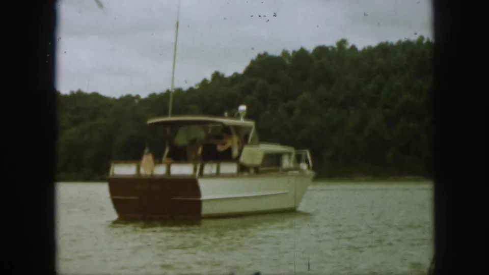 Rear view of the 1961 Michigan lake boat as it moves away from the camera, with passengers under the canopy and trees filling the background.