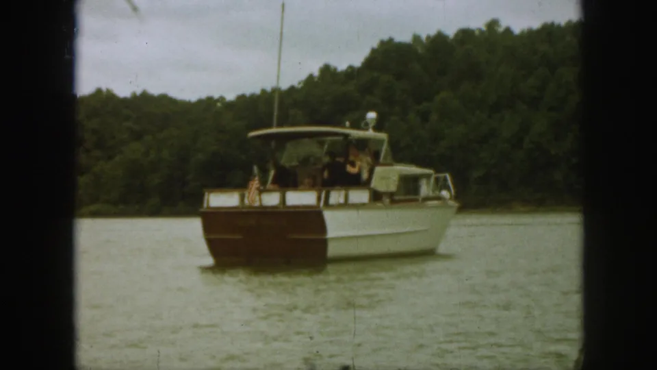A rear three-quarter view shows the motorboat moving farther from the camera on a Michigan lake, with passengers aboard and forested shoreline beyond.