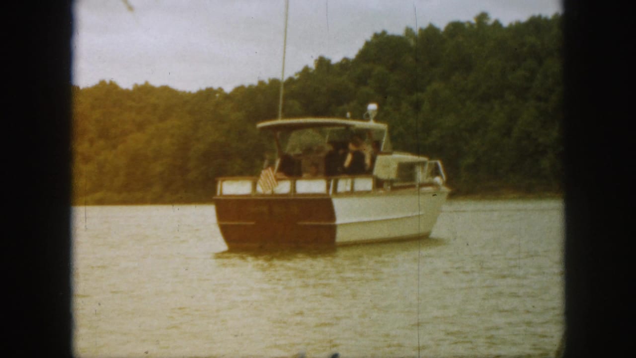 1961 Michigan home-movie frame showing a white and wood tour boat with passengers and a small American flag on a calm lake.
