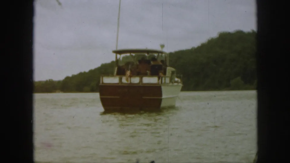Rear view of a Michigan lake tour boat in 1961, with passengers seated beneath a canopy and wooded hills in the background.