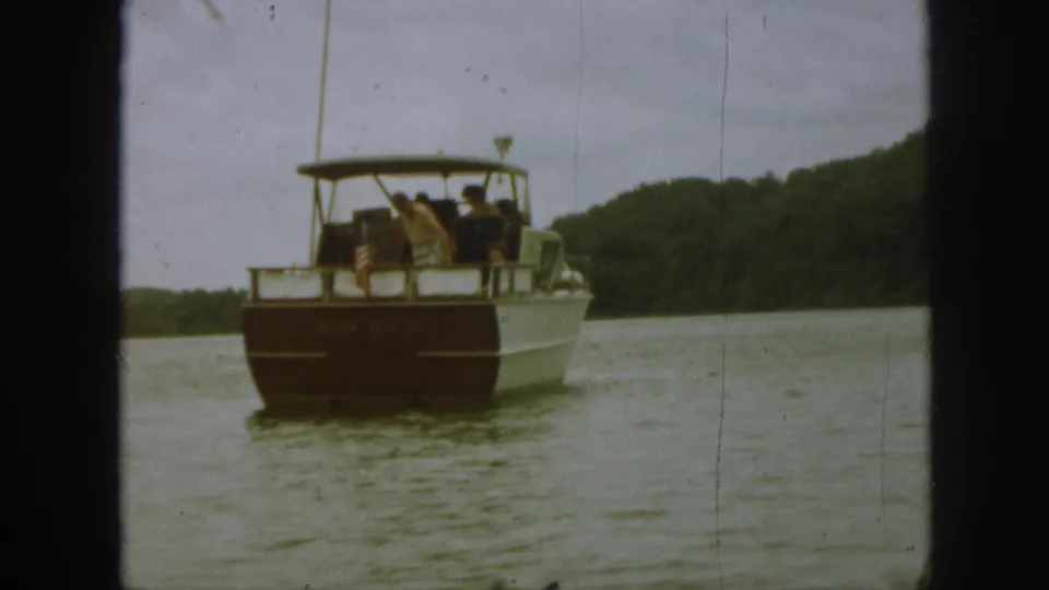 1961 Michigan 8mm frame of a tour boat moving away on calm water, with passengers, canopy, and wooded shoreline visible.