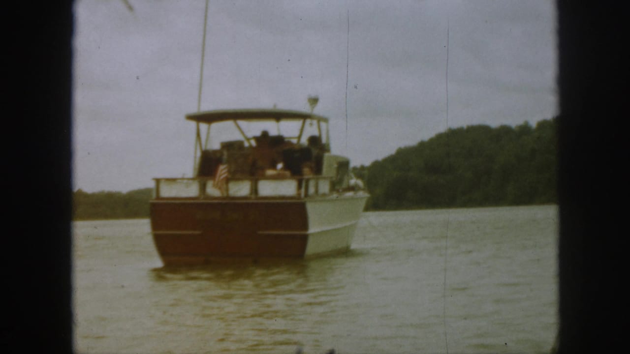 1961 Michigan home-movie frame showing a white-and-brown motorboat on a lake with passengers seated beneath a canopy.