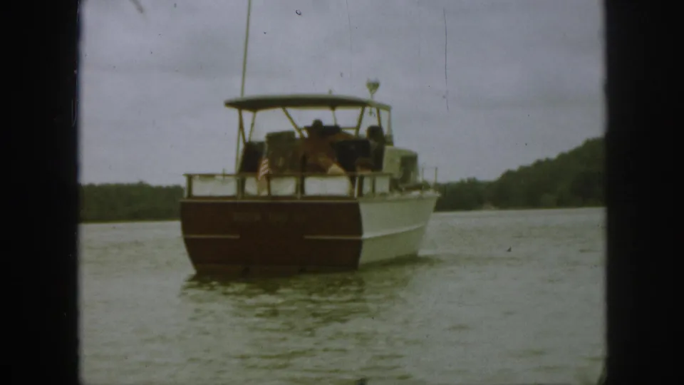 1961 Michigan lake scene with a canopy motorboat carrying several visible people past a wooded shoreline.