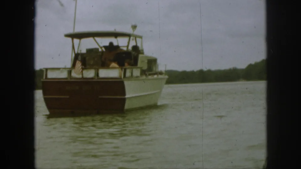 Home-movie view of a 1961 Michigan motorboat crossing a lake, with stern flag, canopy, passengers, and shoreline visible.