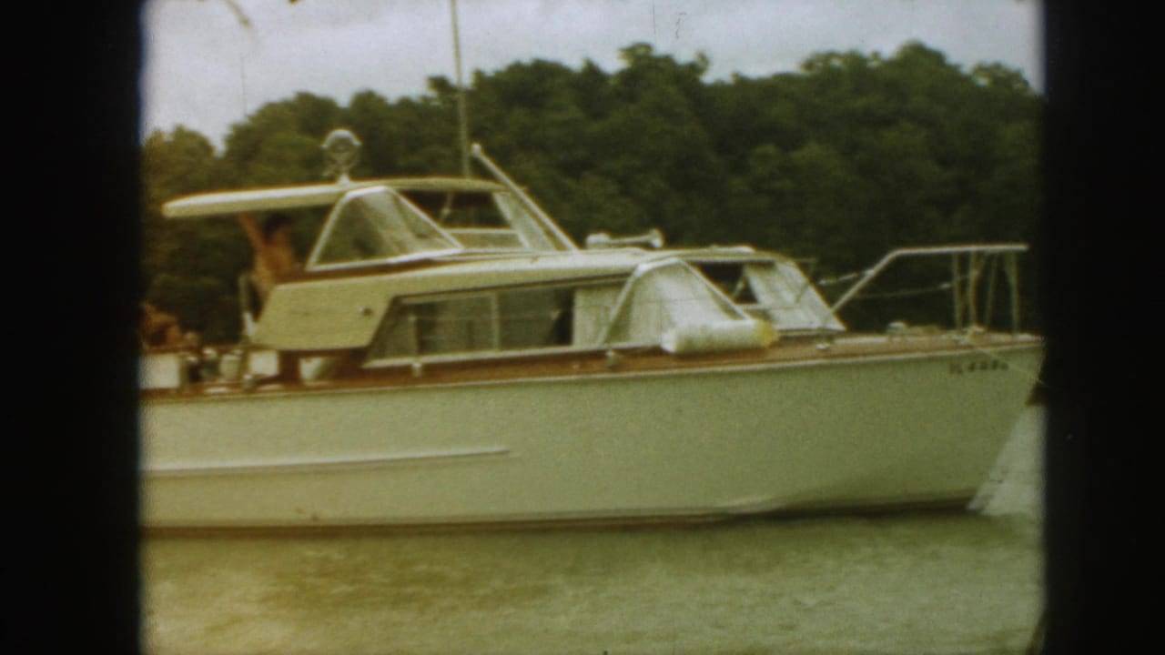 White cabin cruiser on lake water with people on deck and trees behind it.