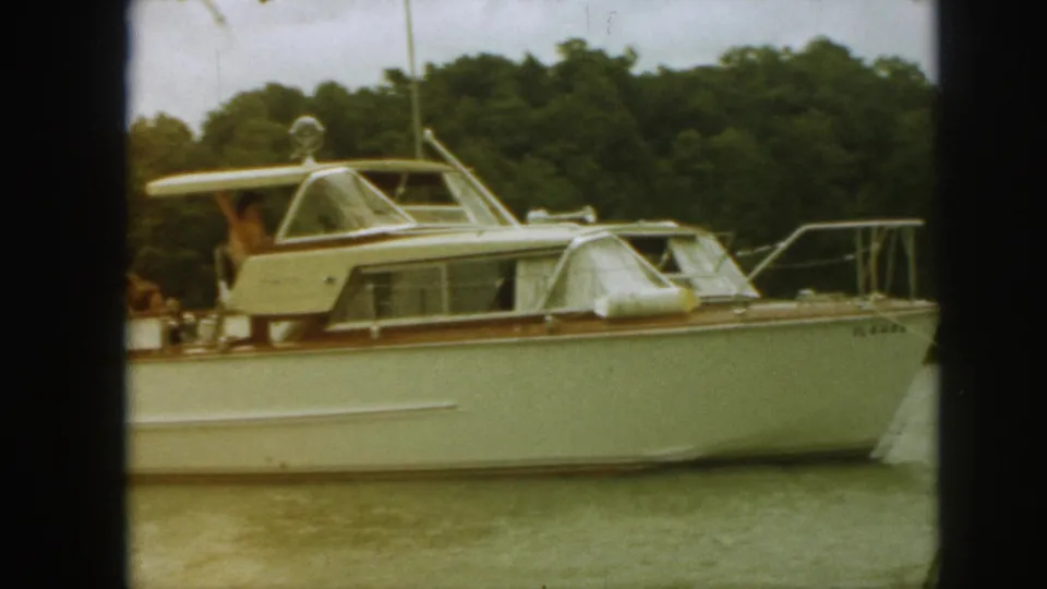 1961 Michigan home-movie frame showing a white motor yacht with wood trim, people on deck, calm water, and a wooded shoreline.