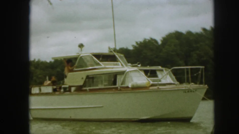 Home-movie frame from 1961 Michigan showing the side of a white yacht, visible passengers, wooded shore, and overcast sky.