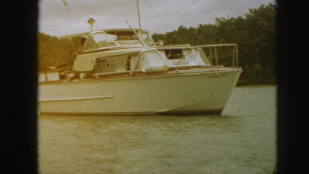 1961 Michigan home-movie frame showing a white motor yacht with cabin, railings, and fenders moving across calm lake water.