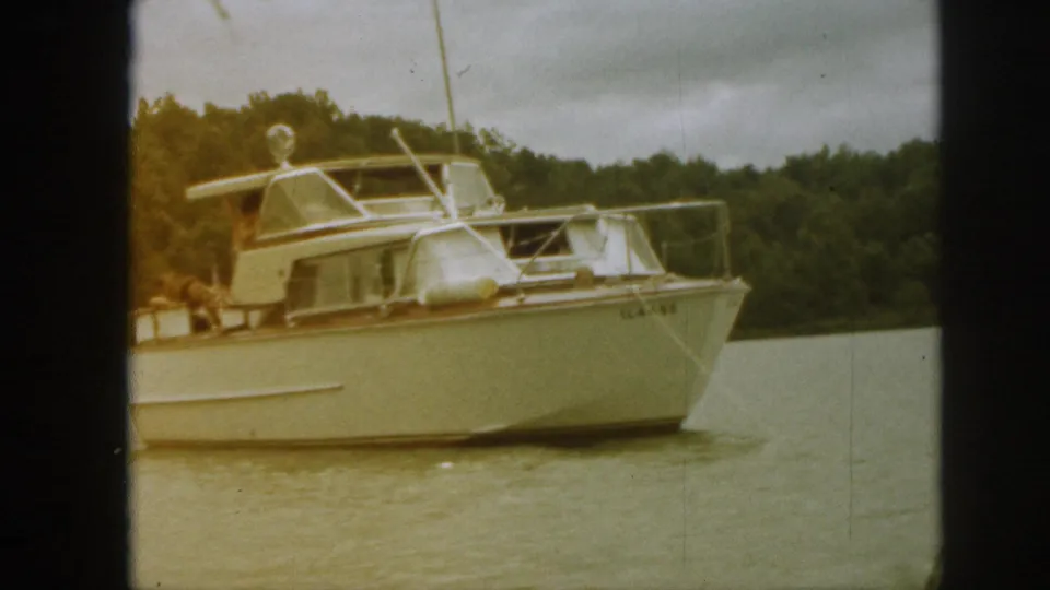 A 1961 Michigan 8mm frame shows the yacht closer in view, with cabin windows, railings, fenders, water, and wooded shore.
