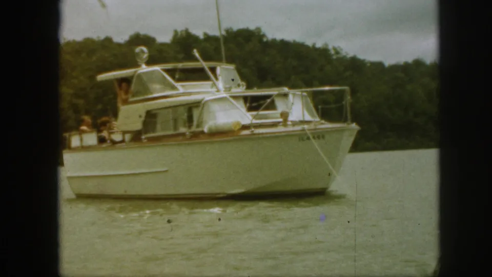 1961 Michigan home-movie frame of a white cabin cruiser on calm water, with people faintly visible near the cabin and wooded shore behind.