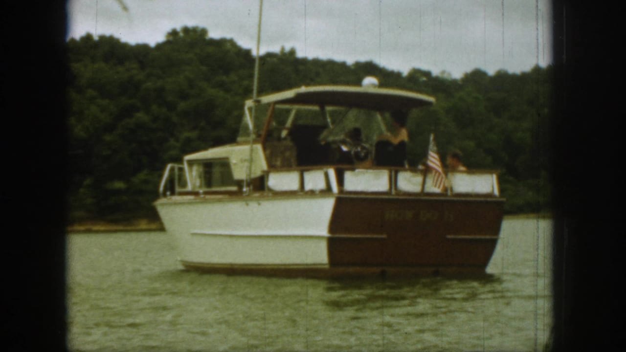 1961 Michigan home-movie frame with a white-and-brown cabin motorboat centered against water and dense shoreline trees.