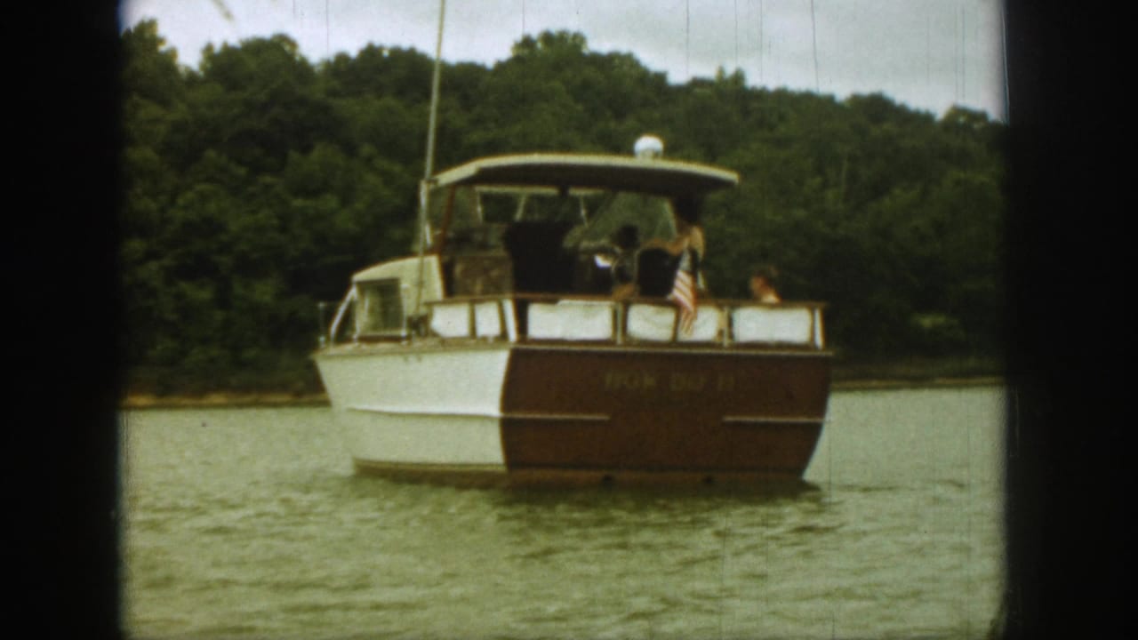 Final 1961 Michigan lake frame showing the motorboat from aft angle, with occupants, U.S. flag, and wooded shore visible.