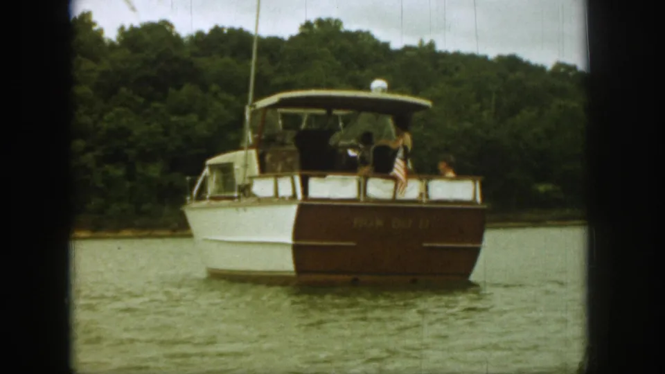 Final 1961 Michigan lake frame showing the motorboat from aft angle, with occupants, U.S. flag, and wooded shore visible.