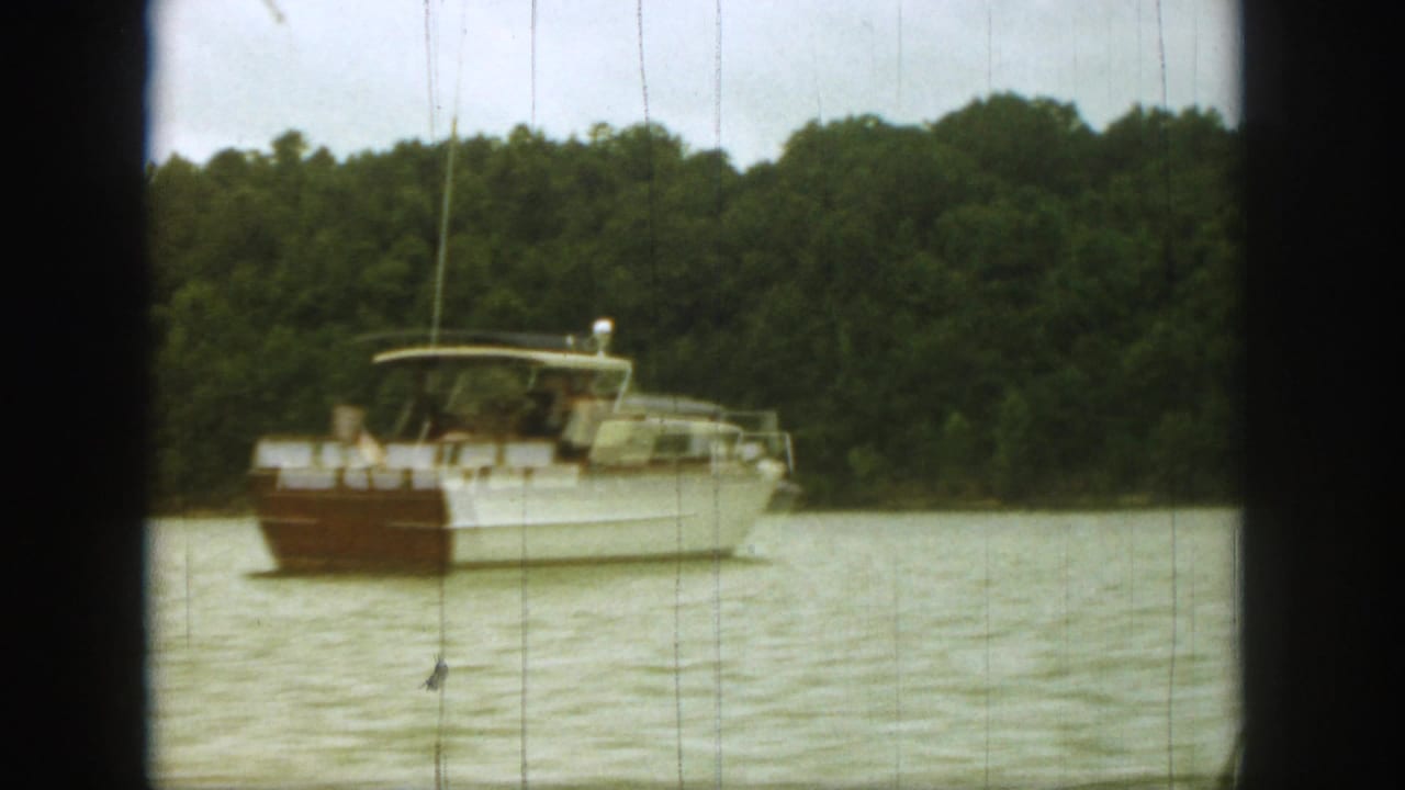 1961 Michigan lake scene with a white and brown motorboat angled across the water near wooded shore.