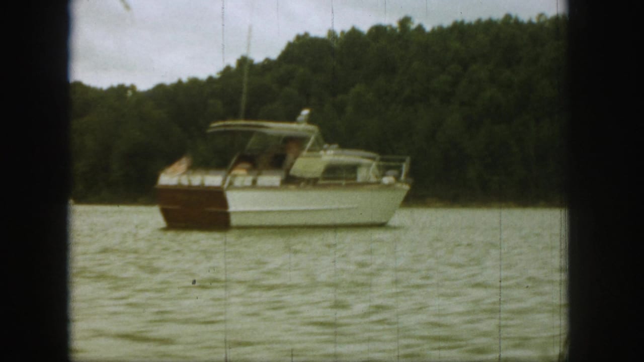 Side view of the classic motorboat on a Michigan lake, with passengers under the cabin roof and trees behind.