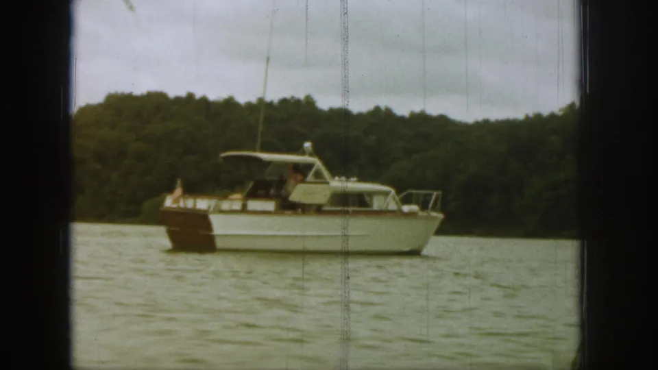 1961 Michigan home-movie frame of a canopied motorboat with a small American flag on calm water before dense trees.