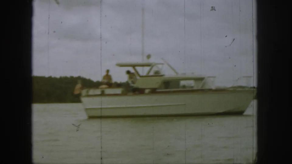 1961 Michigan home-movie frame with a closer side view of the classic motorboat, people aboard, and a low wooded shore behind it.