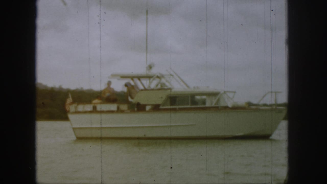 1961 Michigan home-movie frame showing a white motor yacht on calm water with passengers aboard under a cloudy sky.