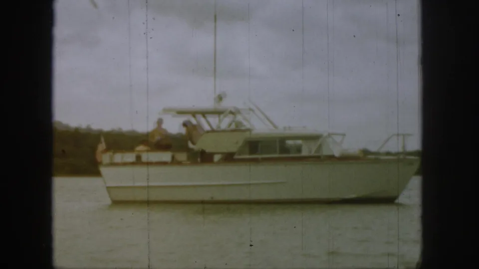 1961 Michigan home-movie frame showing a white motor yacht on calm water with passengers aboard under a cloudy sky.