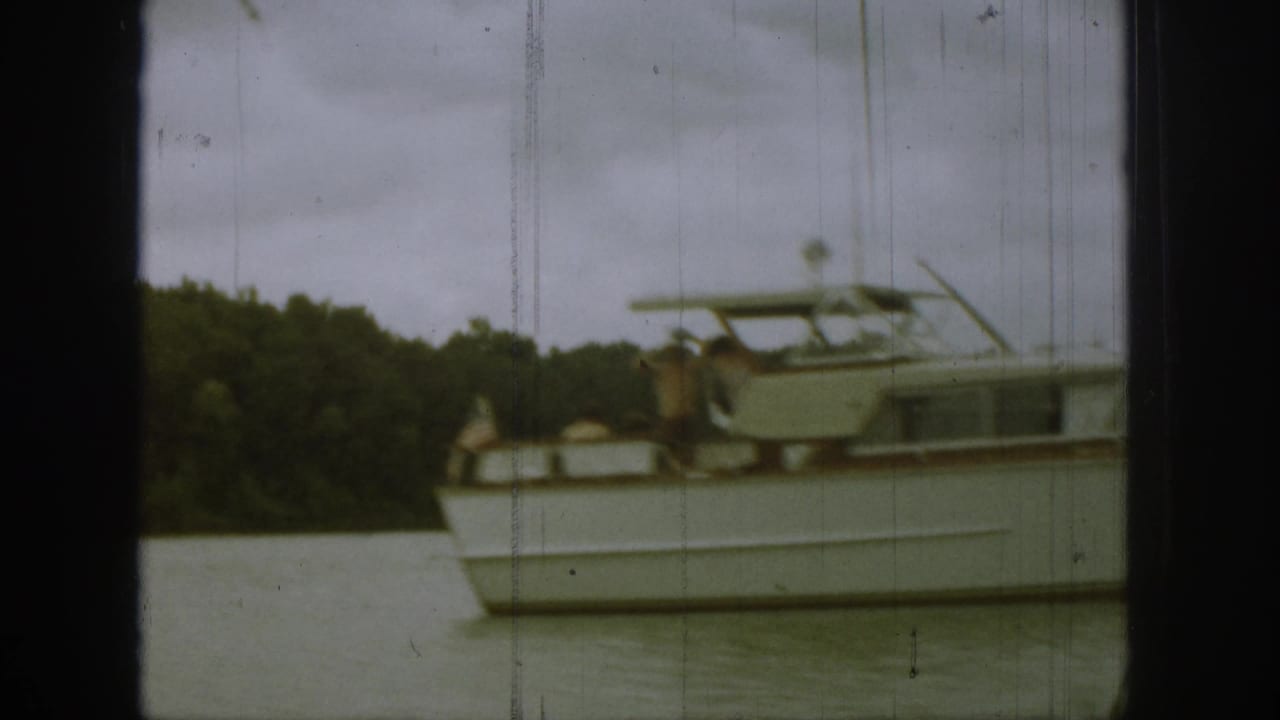 Motor yacht viewed from the side with a passenger near the stern and trees behind.