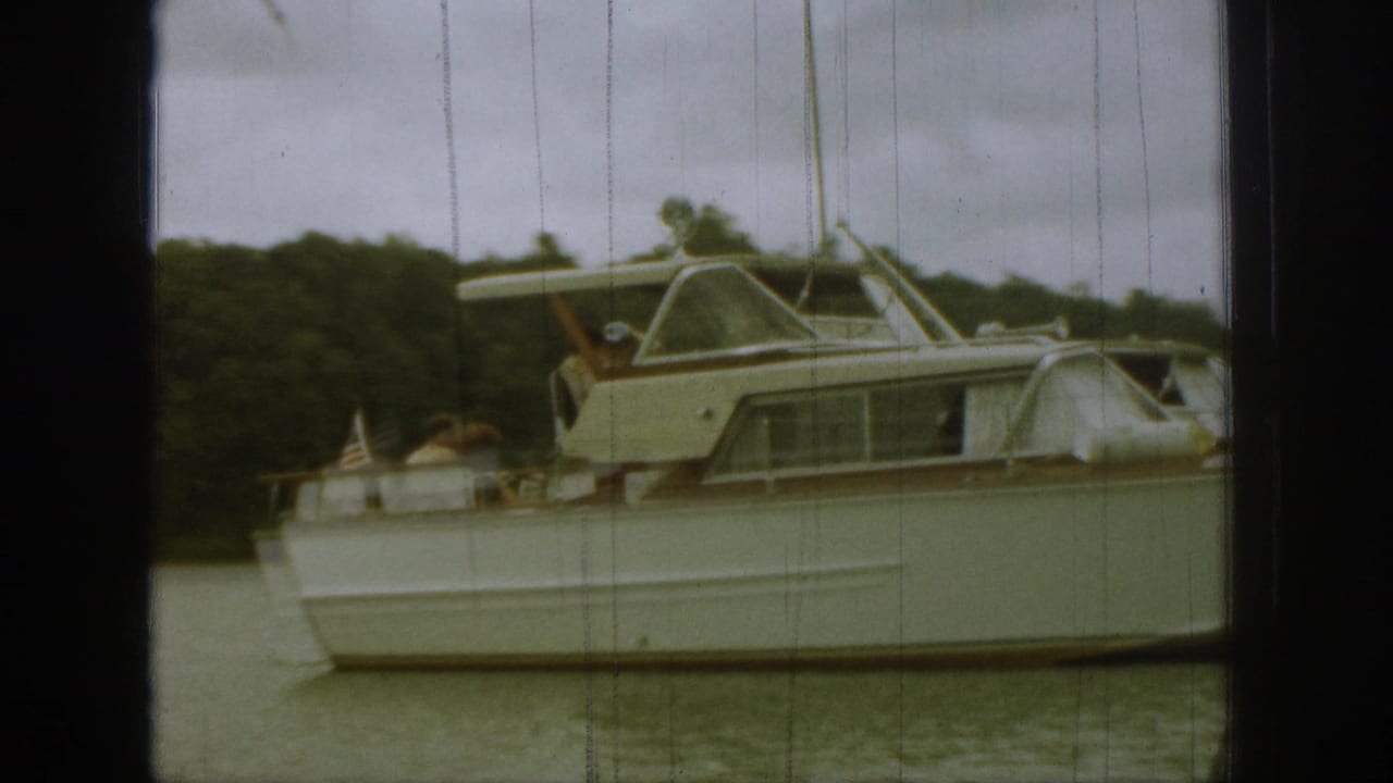 Closer 8mm frame of a white motor yacht, passengers on the stern, and a small American flag near the rear deck.