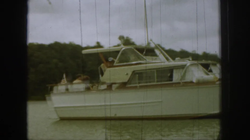 Closer 8mm frame of a white motor yacht, passengers on the stern, and a small American flag near the rear deck.