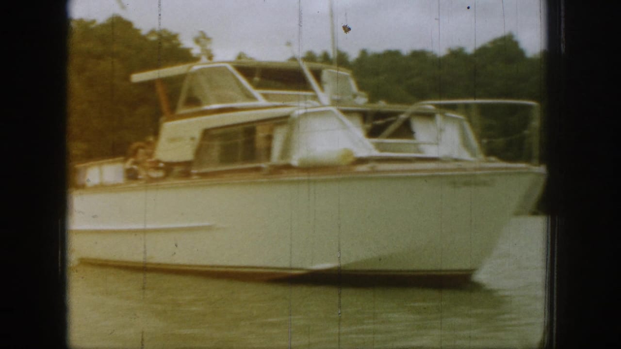Close 1961 Michigan frame of the cabin cruiser’s bow and cabin area, with passengers partly visible and aged film marks throughout.
