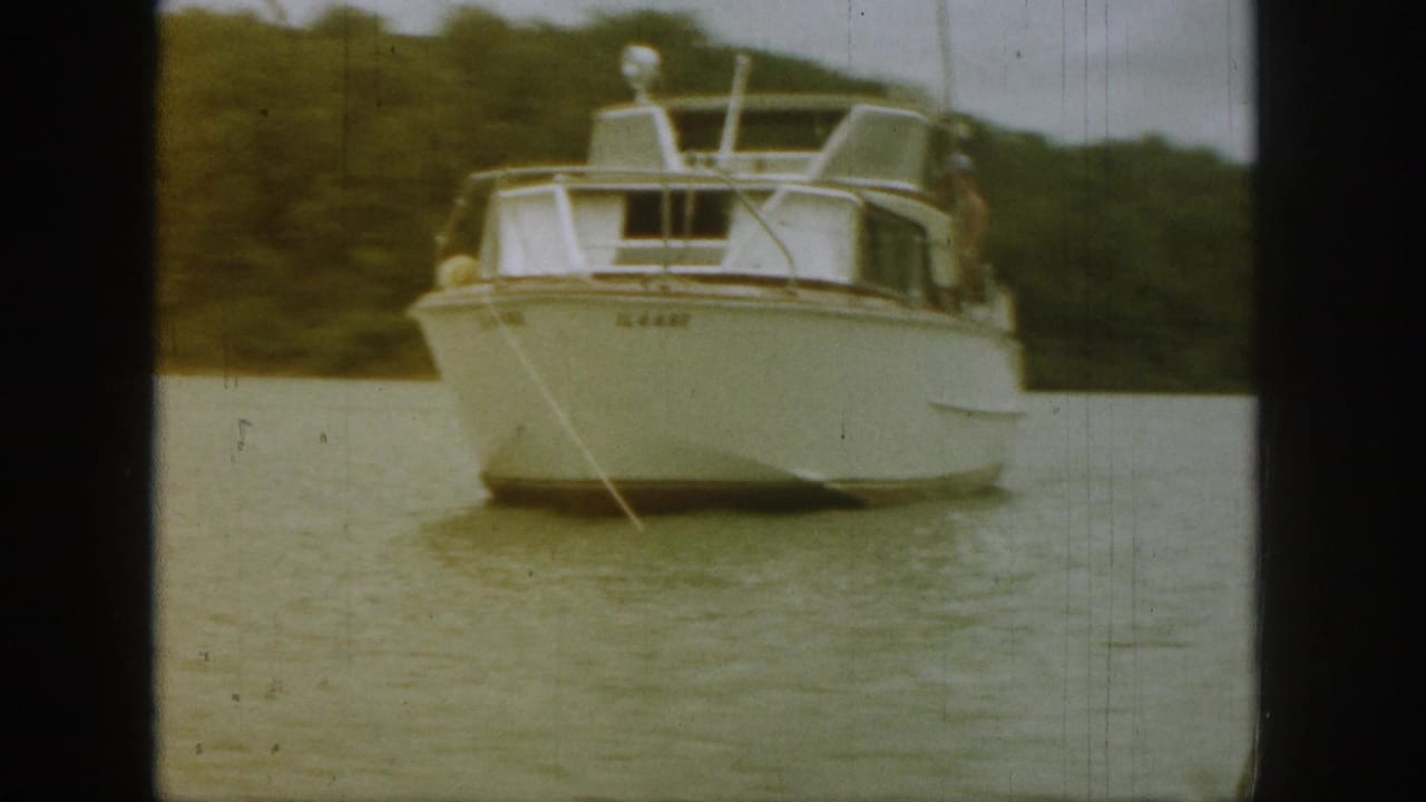 1961 Michigan 8mm frame of a white motorboat angled across calm lake water, with trees behind and a tow line visible.